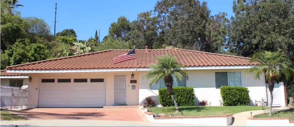 Single-story residential building with a red tiled roof, an American flag mounted near the front door, a two-car garage, and a small front yard with palm trees and bushes under a clear blue sky.