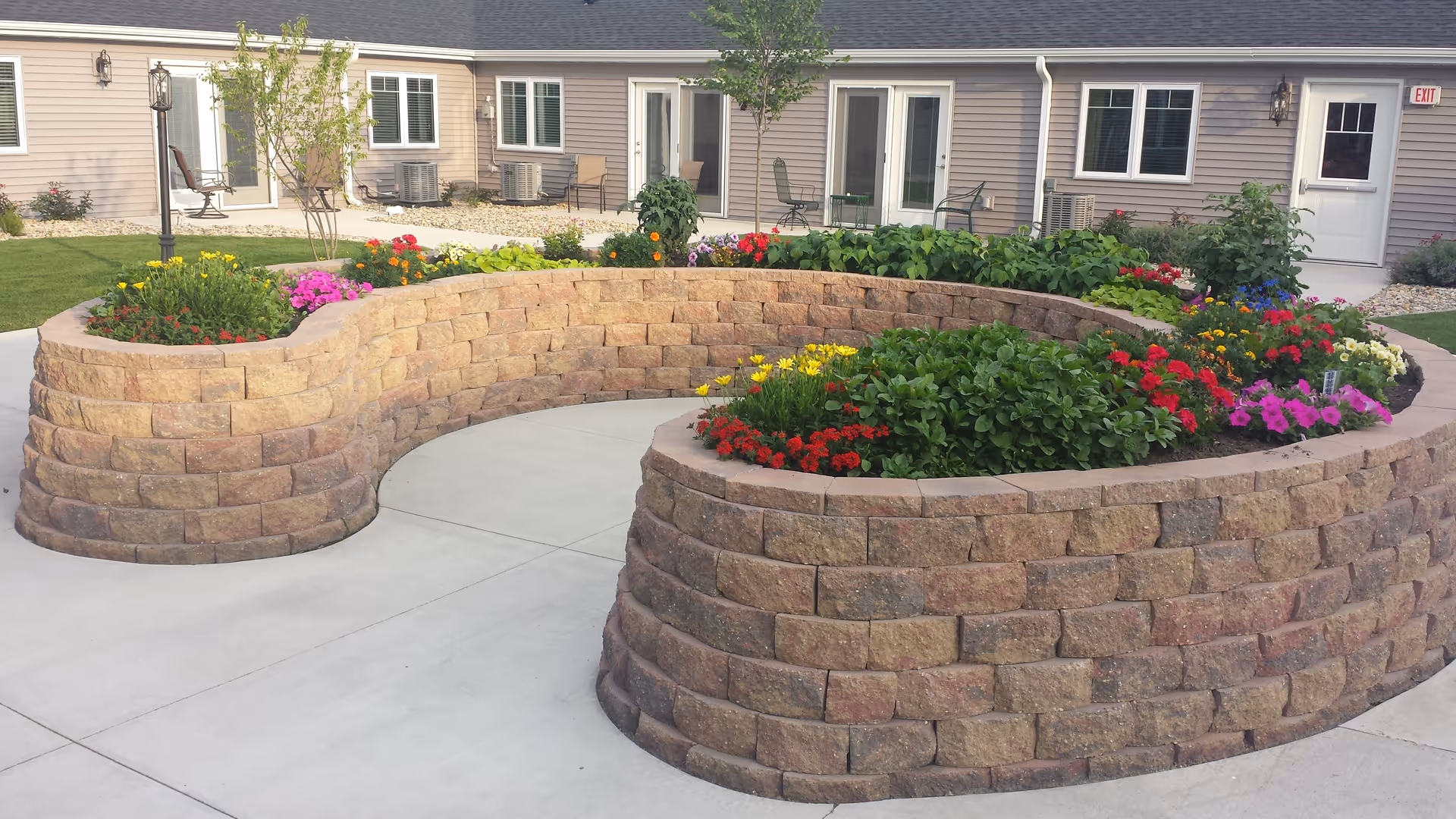 Curved tiered stone planter filled with colorful flowers in a concrete courtyard outside a single-story senior living building with doors and windows.