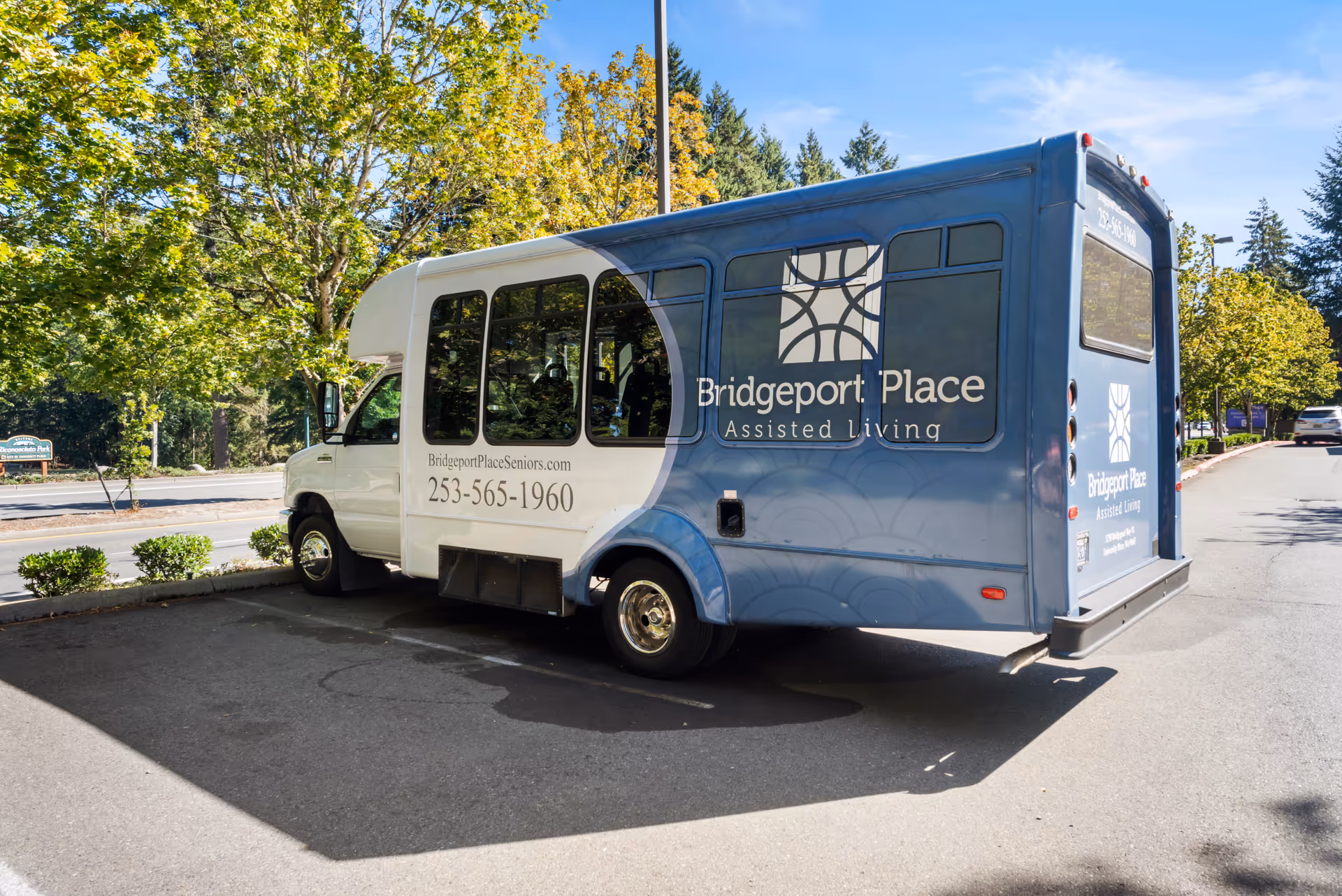 A blue and white shuttle bus parked in a lot with trees and a road in the background. The bus has the logo and text 'Bridgeport Place Assisted Living' along with a phone number and website on its side.