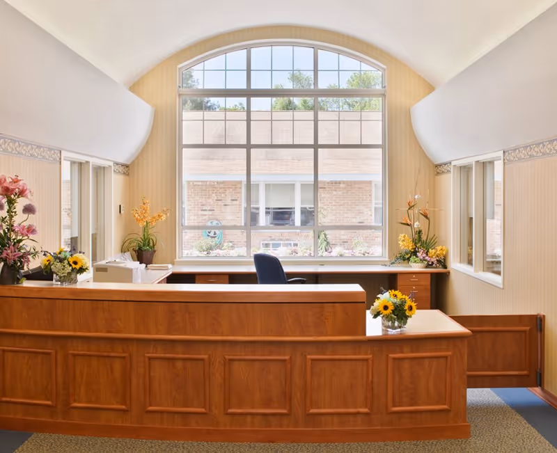 Reception desk area inside a senior living facility with a large arched window, wooden front desk, an office chair, and floral arrangements.