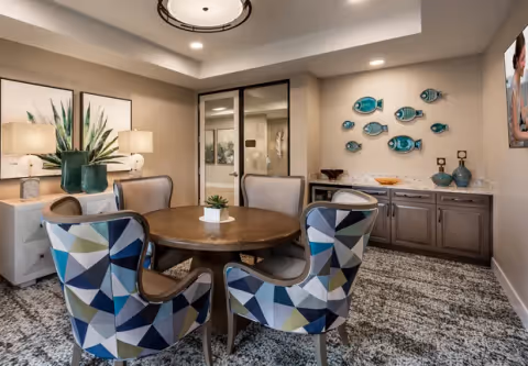 Cozy dining/lounge area with a round wooden table surrounded by patterned upholstered chairs, a sideboard with decorative fish wall art, and soft overhead lighting.