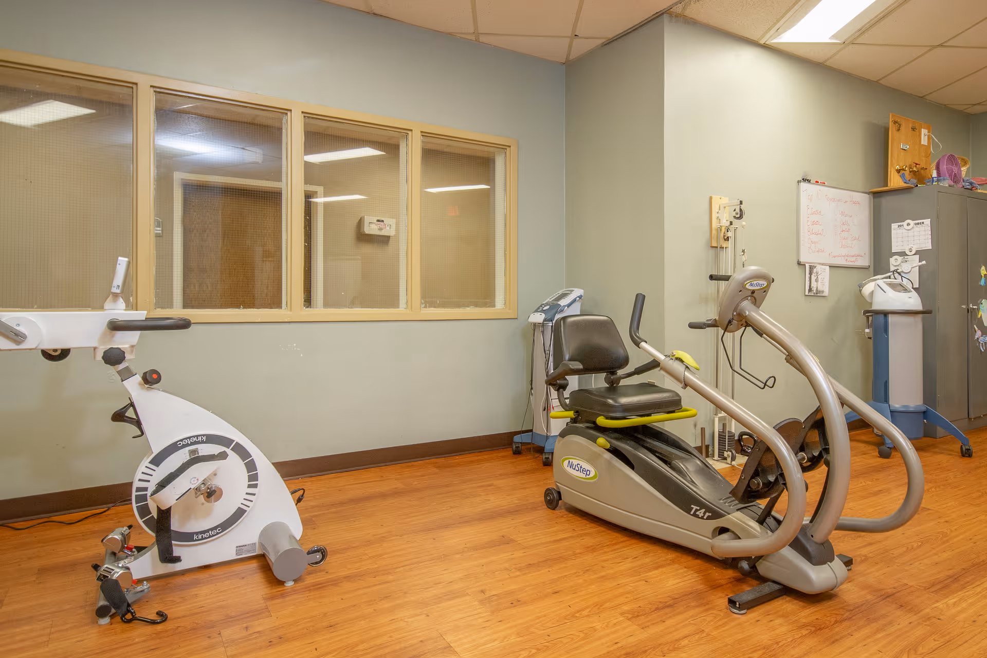 A room with exercise equipment including a white stationary bike and a NuStep T4r recumbent cross trainer on a wooden floor. The walls are light green with a window showing another room, and there is a whiteboard and a cabinet in the background.