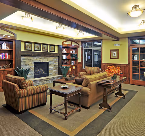 A cozy living room area in a senior living facility with a stone fireplace, wooden built-in shelves, and comfortable seating including a striped armchair and a beige sofa. The room features warm lighting, a wooden coffee table, side tables, and decorative plants and flowers. Large windows and a glass door are visible in the background.