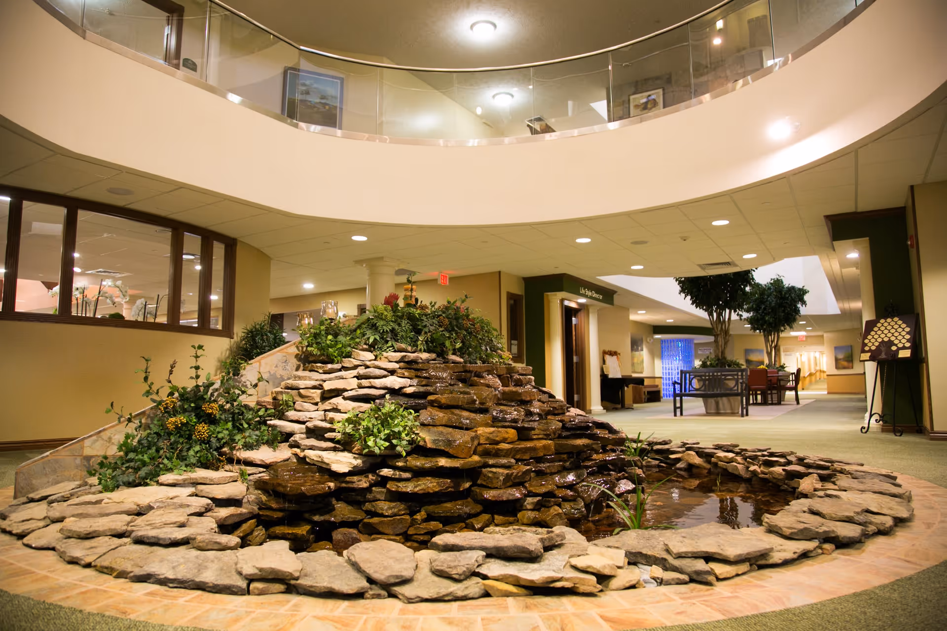 Indoor area of a senior living facility featuring a decorative rock waterfall with plants, surrounded by a circular stone border. The space has a high ceiling with a balcony above, carpeted floors, and seating areas with chairs and tables in the background. The environment is well-lit with ceiling lights and has a calm, welcoming atmosphere.