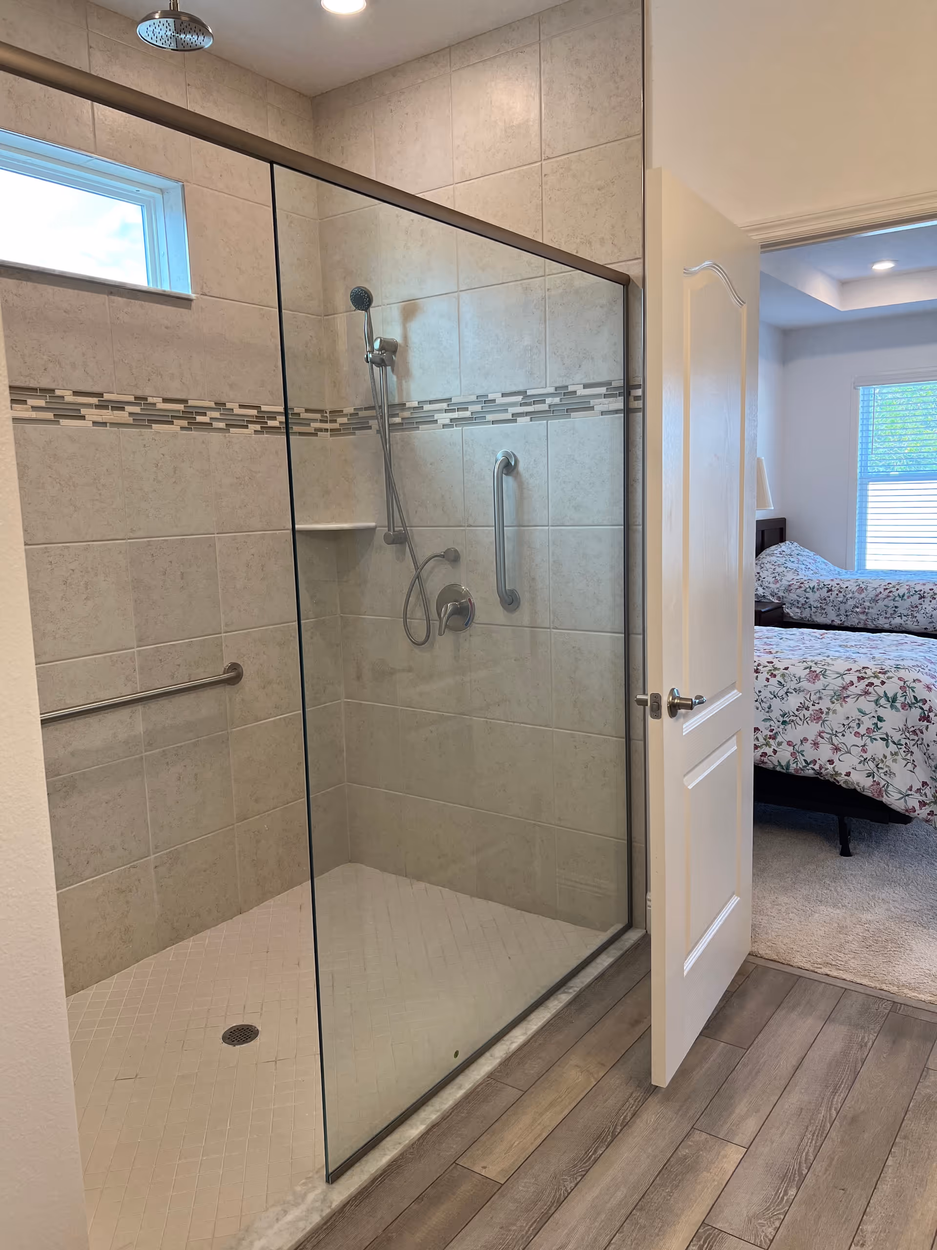 A modern walk-in shower with glass door and tiled walls featuring a decorative horizontal strip. The shower has a handheld showerhead, a rain showerhead, and grab bars for accessibility. The shower floor is tiled with small white tiles. The shower is adjacent to an open door leading to a bedroom with a bed covered in floral bedding and a window with blinds.