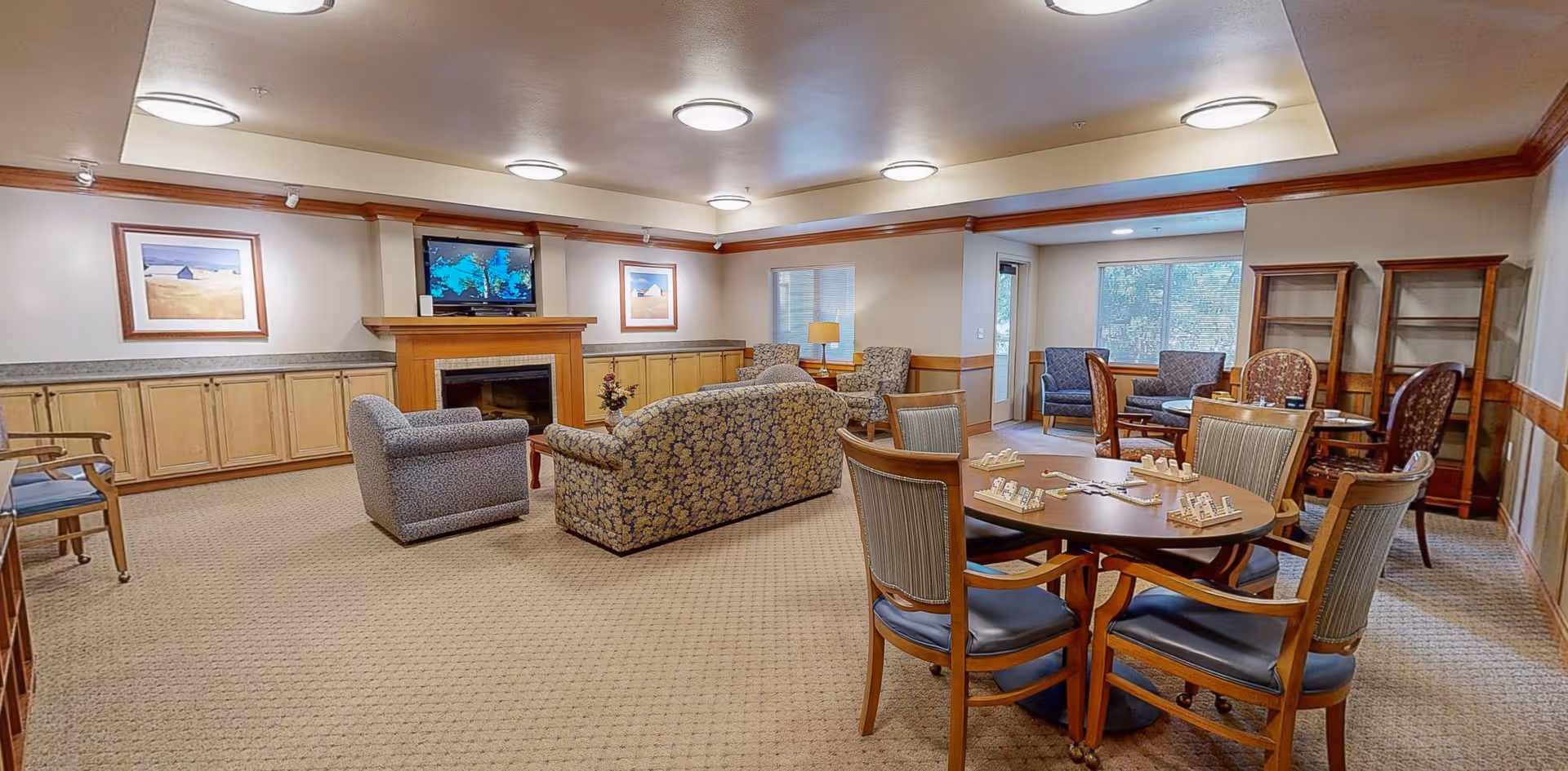 A cozy senior living common area with patterned armchairs and a sofa arranged around a fireplace with a TV mounted above it. There are framed pictures on the walls, a round table with chairs and a domino game set up, and large windows letting in natural light. The room has beige carpet and wood trim accents.