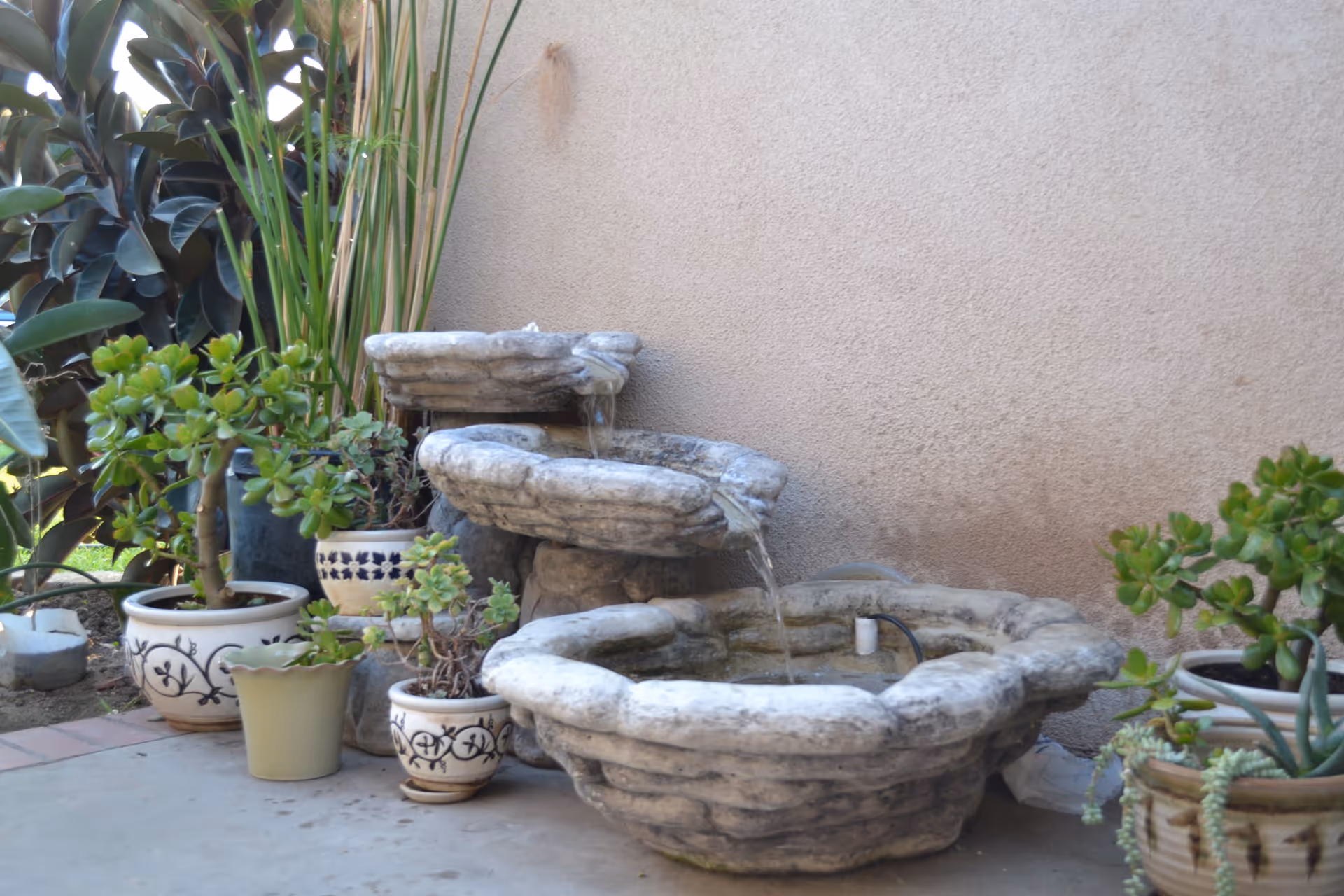 A small outdoor water fountain with three stone basins stacked at different heights, water flowing from the top basin to the bottom. Surrounding the fountain are several potted green plants placed on a concrete surface against a beige stucco wall.