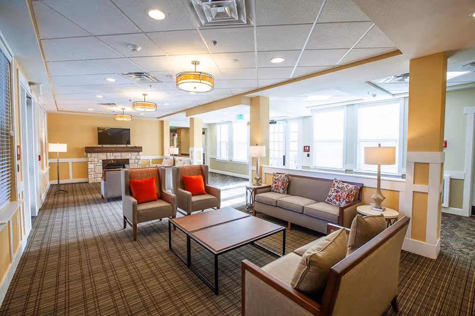 A bright and spacious living room area in a senior living facility with beige walls and carpeted floor. The room features multiple seating options including two armchairs with red cushions, two sofas with patterned pillows, and a wooden coffee table in the center. There are several floor lamps and ceiling lights providing warm lighting. A stone fireplace with a mounted flat-screen TV is visible at the far end of the room. Large windows allow natural light to fill the space.