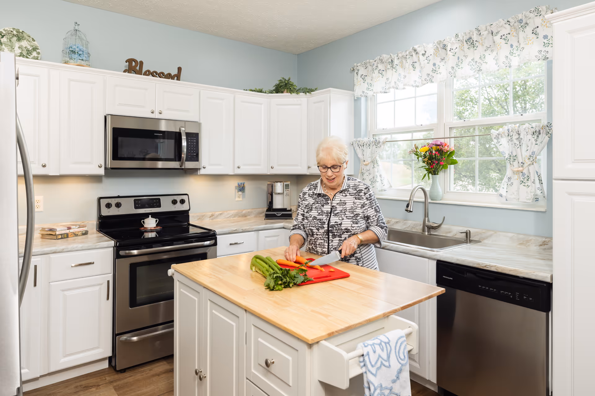 An elderly woman with short white hair and glasses is chopping vegetables on a red cutting board placed on a wooden kitchen island. The kitchen has white cabinets, a stainless steel stove, microwave, and dishwasher. There is a large window with floral curtains letting in natural light, and a vase with flowers on the windowsill.