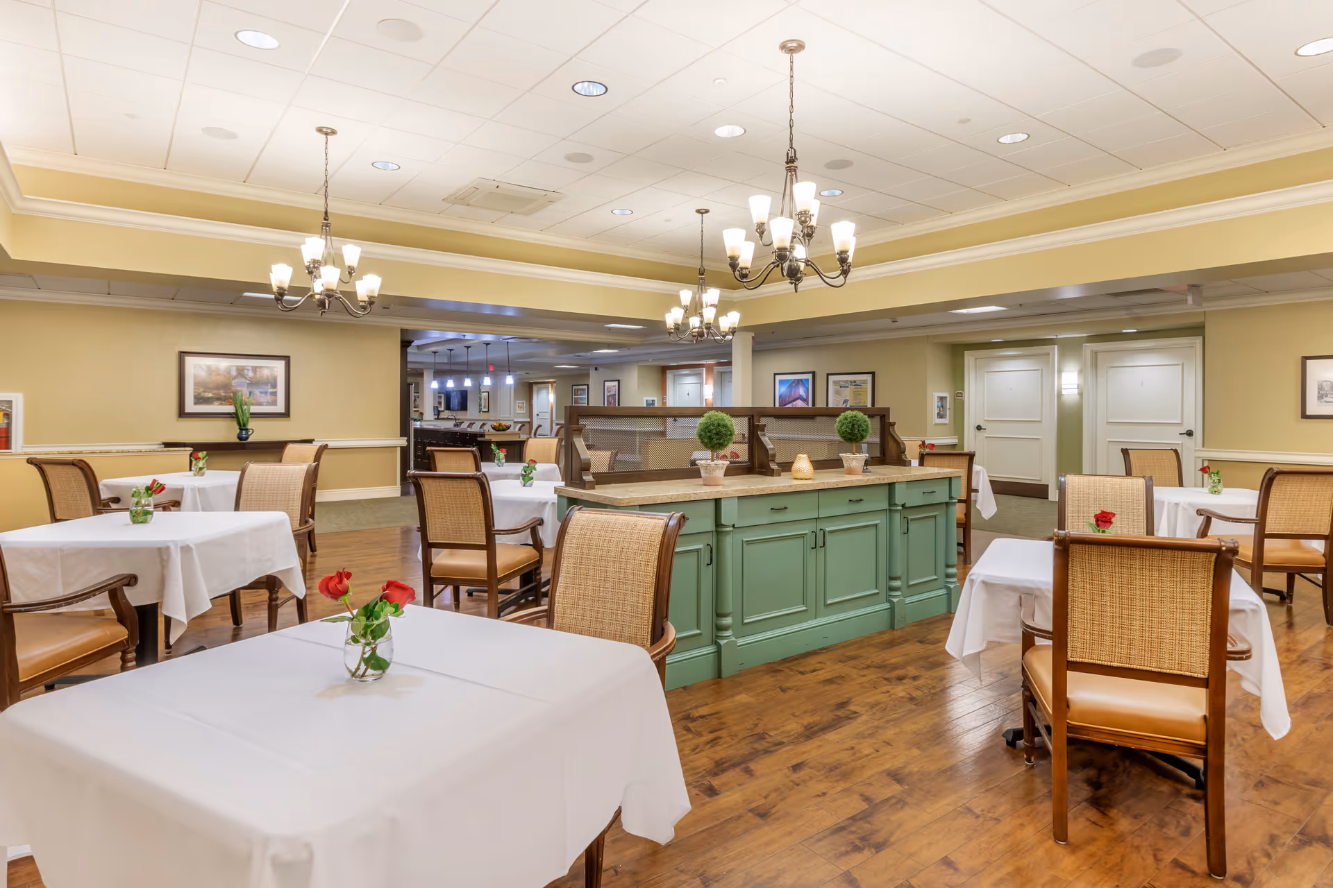 A well-lit dining room with several tables covered in white tablecloths, each adorned with a small vase holding a red rose. The room features wooden chairs with woven backs and leather seats, a green cabinet in the center with decorative plants, chandeliers hanging from the ceiling, and framed artwork on the beige walls.