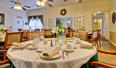Round dining table set with plates, glasses, and napkins in a bright senior living dining room.