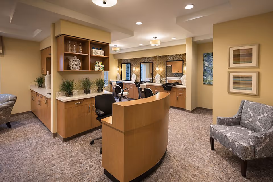 Interior view of a salon area in a senior living facility with a curved wooden reception desk, salon chairs, mirrors, and cabinets. The space is decorated with plants, vases, and framed artwork on the walls. There is a patterned armchair on the right side.