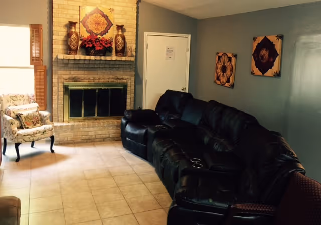 A cozy living room with a black leather sectional sofa featuring cup holders, a floral upholstered armchair near a window with wooden blinds, a brick fireplace with decorative vases and a floral arrangement on the mantel, and two framed floral artworks on a gray wall.