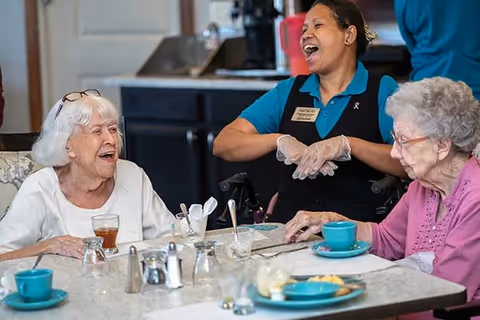 Two elderly women sitting at a dining table with cups and plates, smiling and engaging with a cheerful staff member wearing gloves and a name tag in a kitchen or dining area.