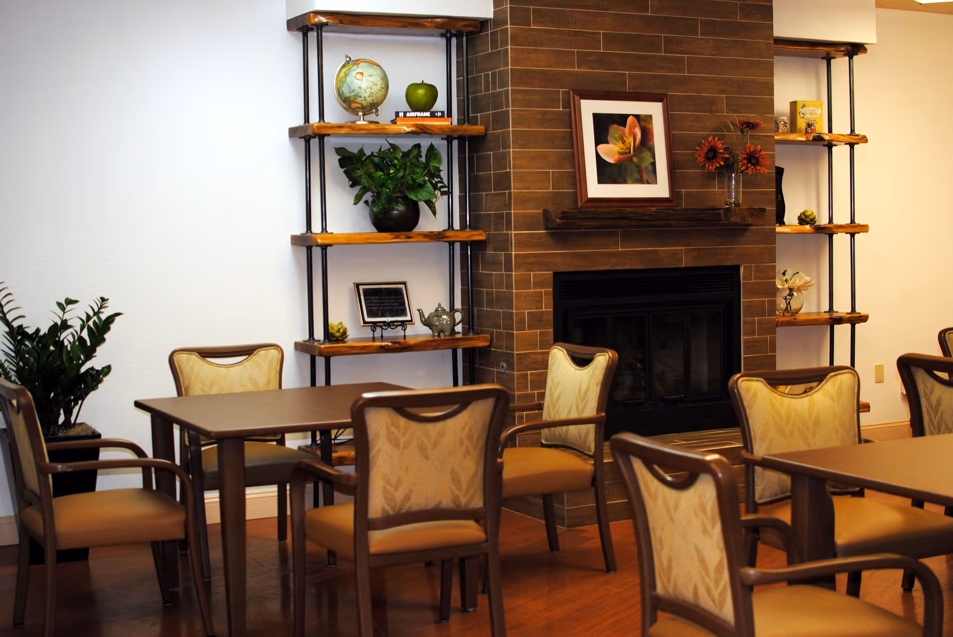 A cozy interior room with several wooden tables and cushioned chairs arranged around them. There is a brick fireplace in the background with a framed flower picture above it. On either side of the fireplace are wooden shelves holding decorative items such as a globe, plants, and small ornaments. The room has a warm and inviting atmosphere.