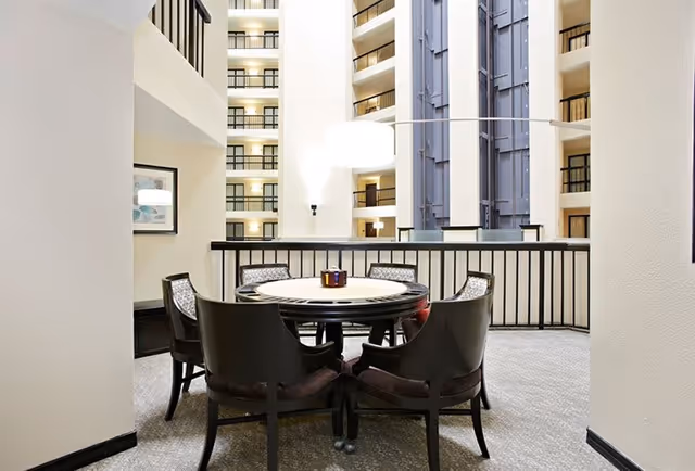 A seating area with a round table and six chairs arranged around it, located in an indoor atrium space with multiple floors and balconies visible in the background. The area is carpeted and has a modern, clean design with neutral colors.
