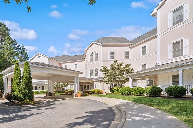 Exterior view of HeartFields at Cary senior living facility showing a multi-story building with beige walls and white trim, a covered driveway entrance, well-maintained landscaping with bushes and trees, and a clear blue sky.