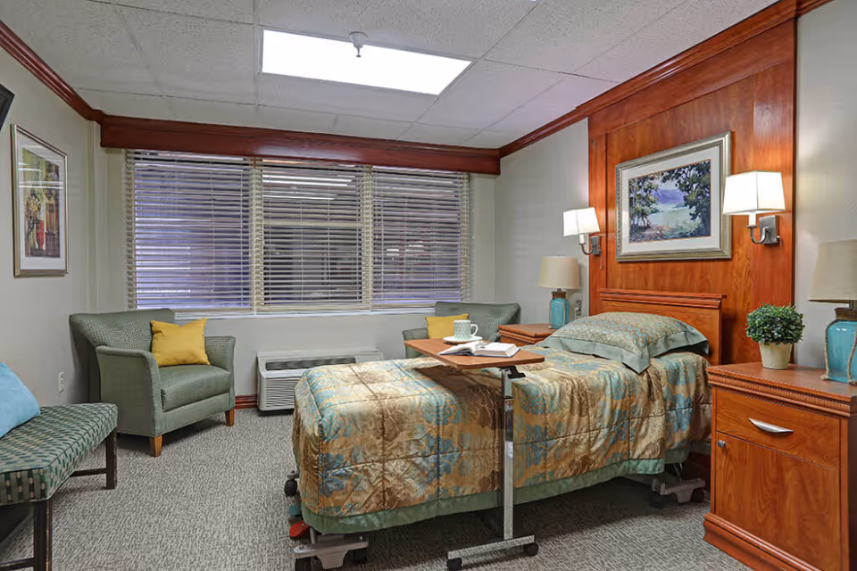 A well-lit bedroom in a senior living facility featuring a single hospital-style bed with patterned bedding, a wooden headboard with two wall-mounted lamps, a bedside table with a lamp and a small potted plant, a green armchair with a yellow pillow, a bench, and a large window with blinds.