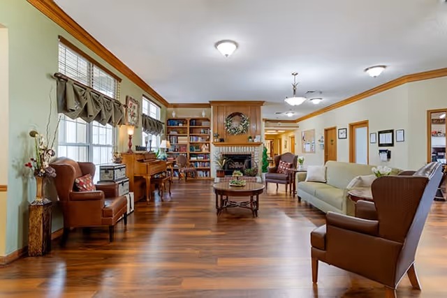 A spacious and well-lit living room area in a senior living facility with wooden flooring, a central fireplace decorated with a wreath, bookshelves, a piano, and various comfortable seating including armchairs and sofas arranged around a coffee table.