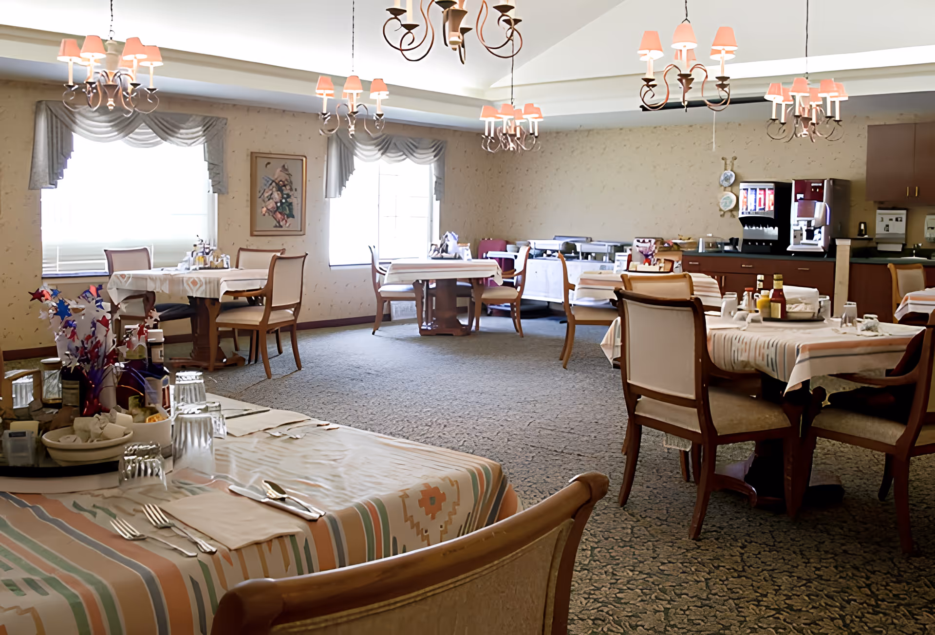 A dining room in Cheney Care Center with multiple tables covered in patterned tablecloths, set with utensils, glasses, and condiments. The room has carpeted floors, large windows with draped curtains, chandeliers with pink lampshades hanging from the ceiling, and a beverage station with a soda fountain and coffee machine in the background.