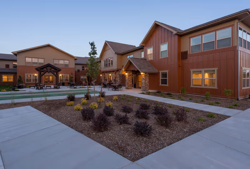 Exterior view of a senior living facility at dusk with warm lights glowing from the windows. The building has a modern design with brown and tan siding, stone accents around the entrances, and a landscaped courtyard with small shrubs and young trees. Concrete walkways surround the garden area.