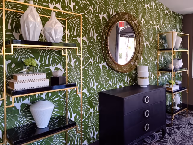 Interior space with green and white leaf-patterned wallpaper, featuring two gold-framed black shelves holding decorative white vases and boxes. A black chest of drawers with circular handles is positioned between the shelves, topped with a white vase. Above the chest is a round, ornate mirror reflecting part of the room and an exit sign.
