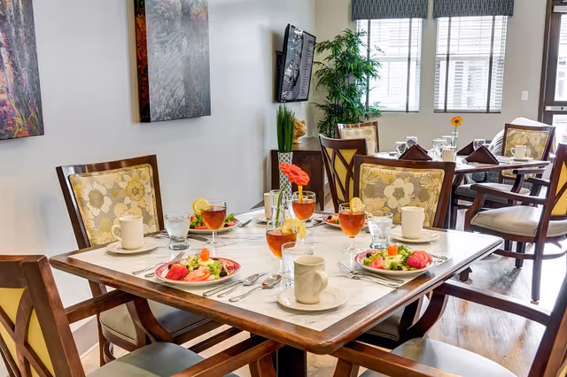 A dining room with wooden tables and chairs set for a meal. Each table has plates with salads, glasses of iced tea with lemon, coffee cups, and silverware. The room has large windows with blinds, a wall-mounted TV, and decorative plants and artwork on the walls.