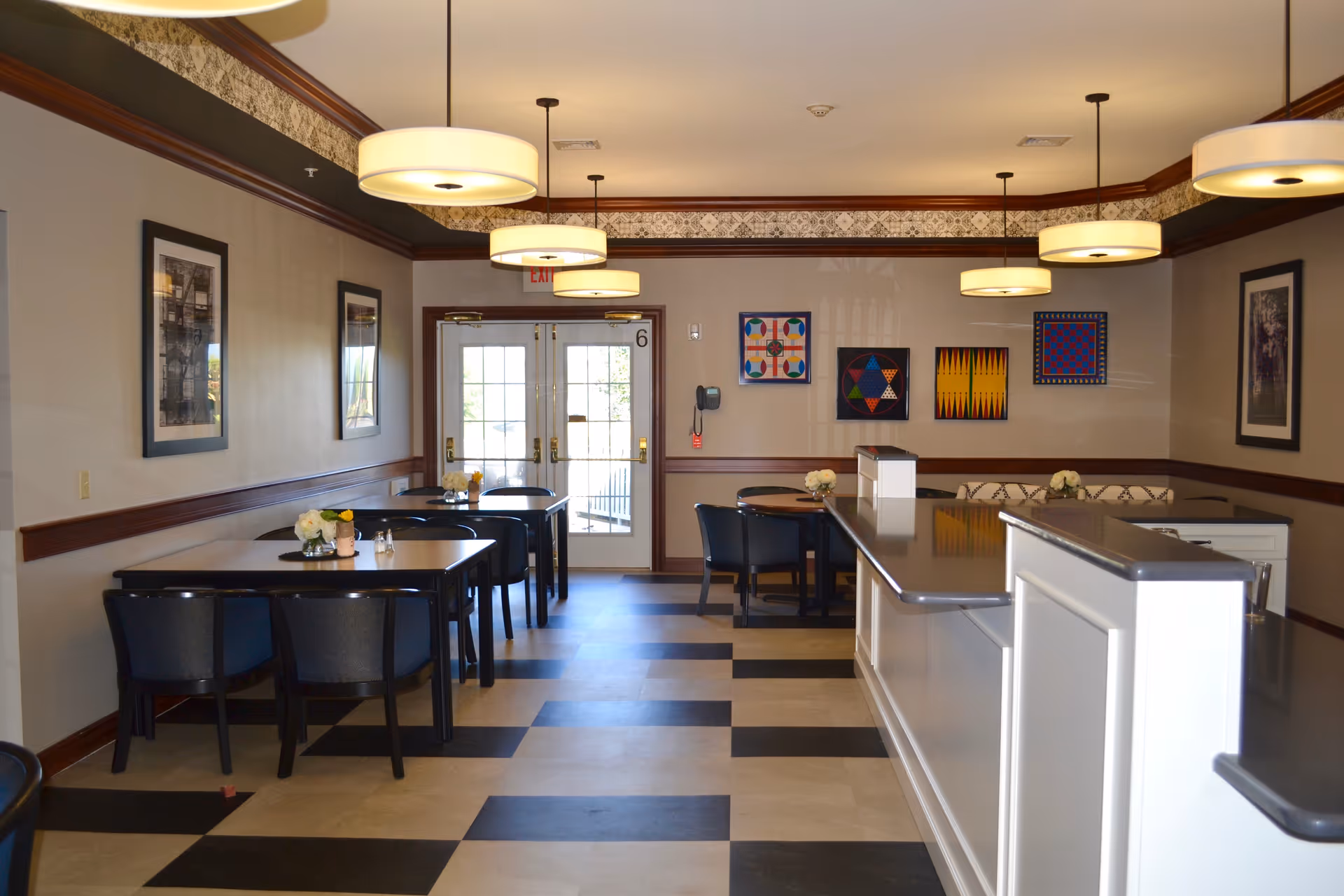 Interior view of a dining area in a senior living facility with several tables and chairs arranged neatly. The room features a checkered floor pattern, pendant lights hanging from the ceiling, framed artwork on the walls, and a counter on the right side. Double glass doors are visible at the far end of the room.