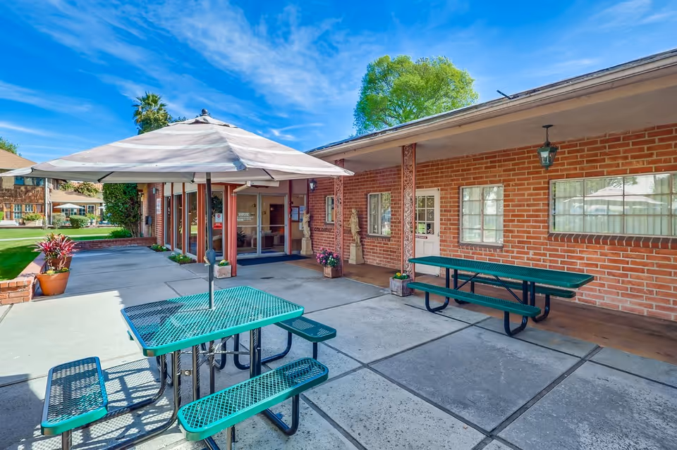 Outdoor patio area at Colonial Gardens Nursing Home with green metal picnic tables, one with a large beige umbrella. The patio is paved with concrete and adjacent to a brick building with windows and a door. There are potted plants and a green lawn with trees and other buildings in the background under a blue sky with some clouds.