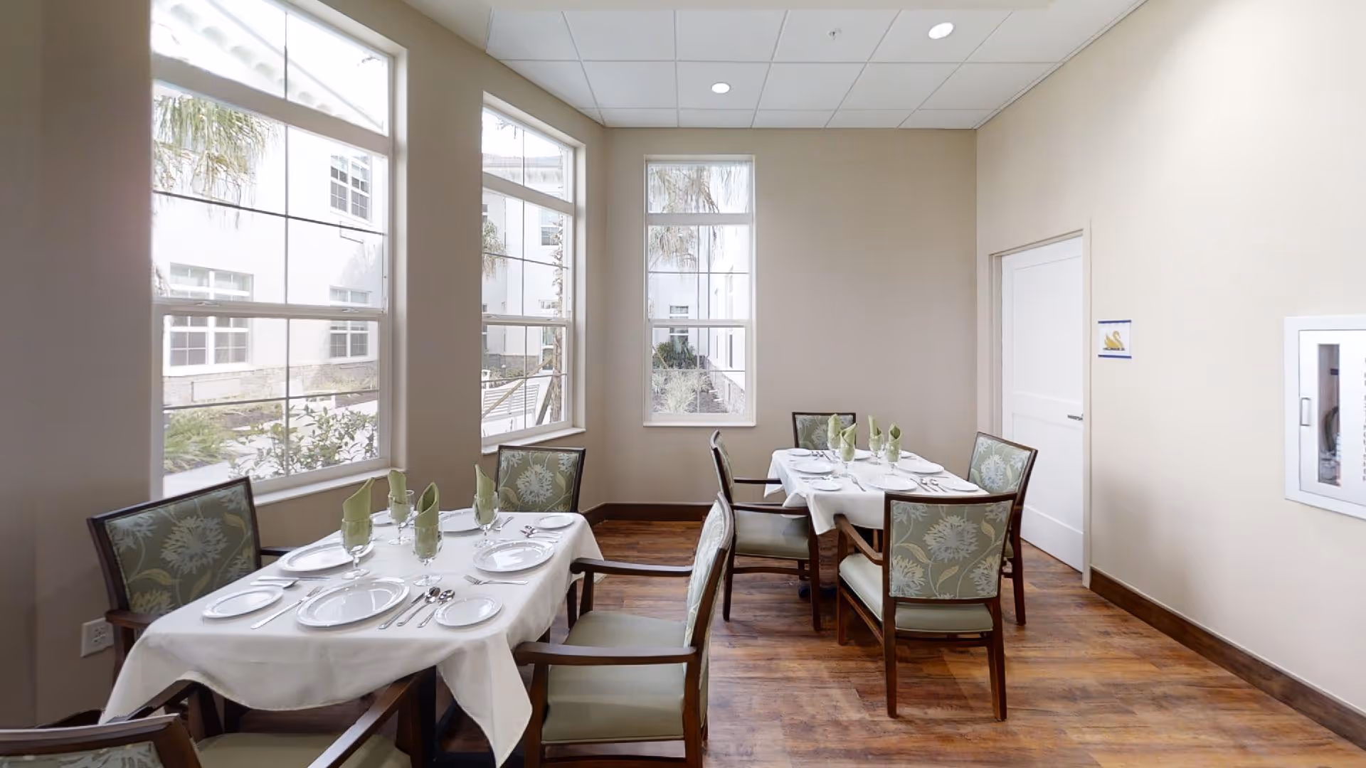 A bright dining room with two tables set for meals, each covered with white tablecloths and neatly arranged with plates, glasses, silverware, and green folded napkins. The room has large windows letting in natural light and wooden flooring, with beige walls and floral upholstered chairs.