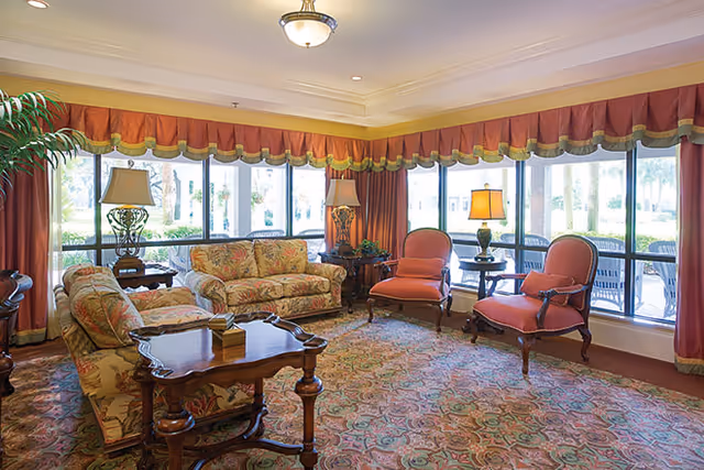 Sunny seating area with floral sofas, upholstered armchairs, wooden side tables and lamps in front of large windows overlooking greenery.