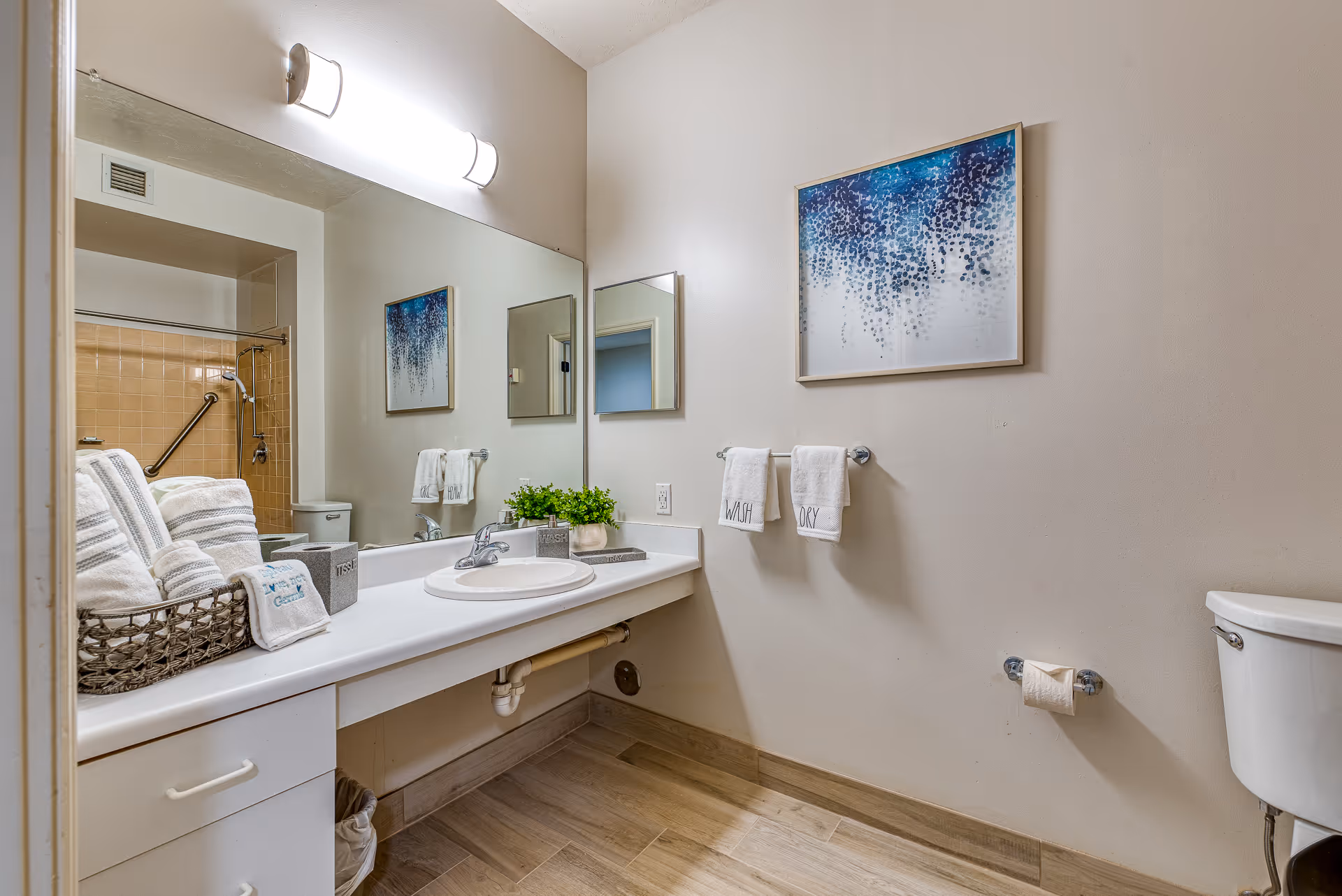 A clean and well-lit bathroom featuring a white countertop with a sink, a large mirror, and a basket filled with neatly folded towels. On the wall, there is a blue and white abstract painting and two hand towels hanging on a rack. The bathroom also includes a toilet and a shower area with beige tiles and a grab bar.