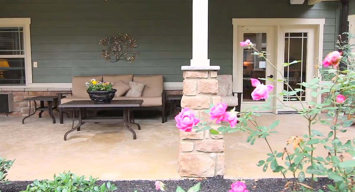 Outdoor patio area with a cushioned bench, two side tables, a coffee table with a flower pot, and a stone pillar. Pink flowers and green plants are in the foreground, and a green exterior wall with a window and glass door is in the background.