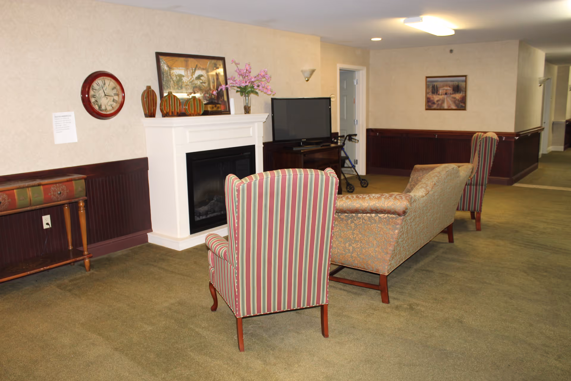 A cozy common area in a retirement community featuring a striped armchair, a patterned sofa, a white fireplace with decorative pumpkins and a flower vase on the mantel, a wall clock, a flat-screen TV on a stand, and a walker nearby. The room has green carpet, beige walls with dark wood paneling, and framed artwork on the walls.