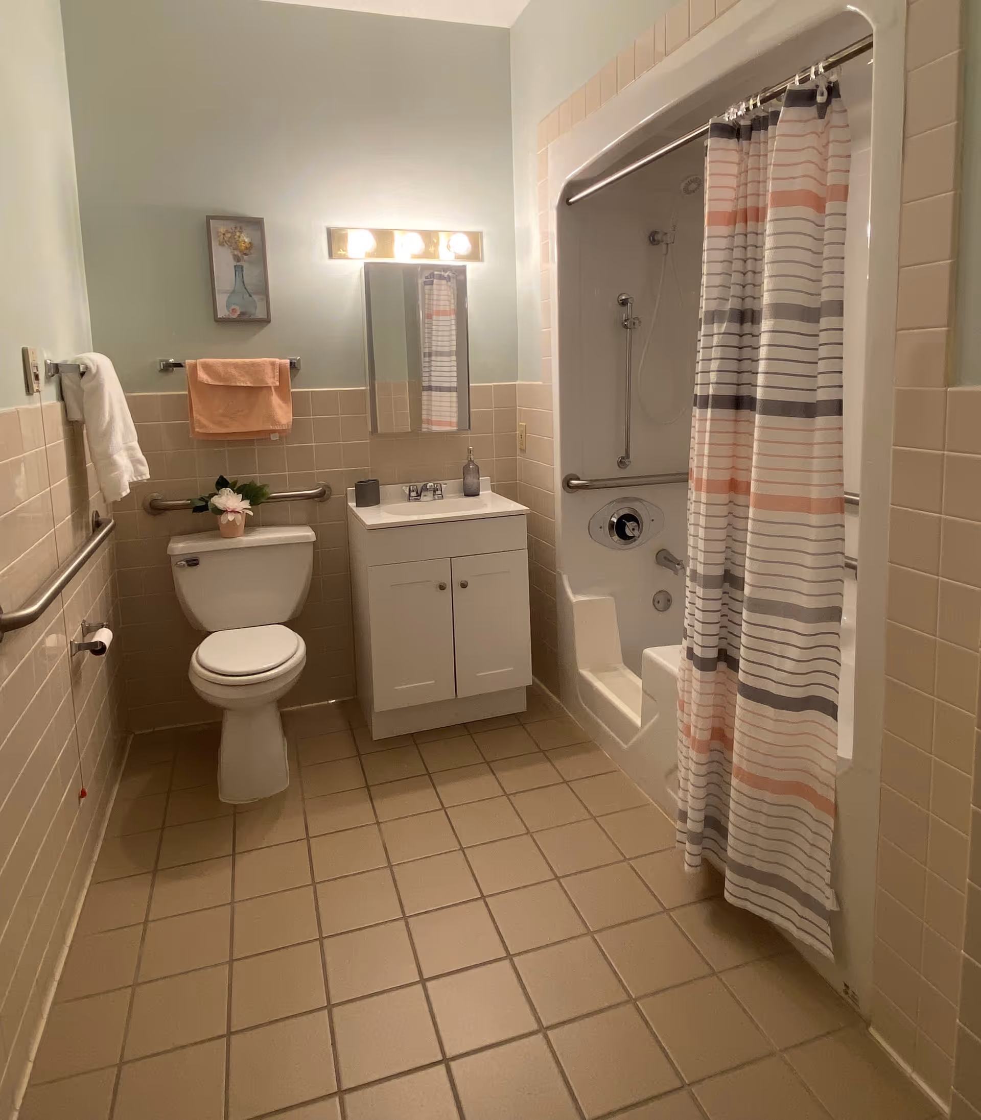 A clean and accessible bathroom with beige tiled floor and walls halfway up. It features a white toilet with a grab bar on the wall behind it, a white vanity with a sink and a mirror above it, and a bathtub with a built-in seat and a striped shower curtain in shades of gray, white, and peach. There are towels hanging on the wall and a small framed picture above the toilet.