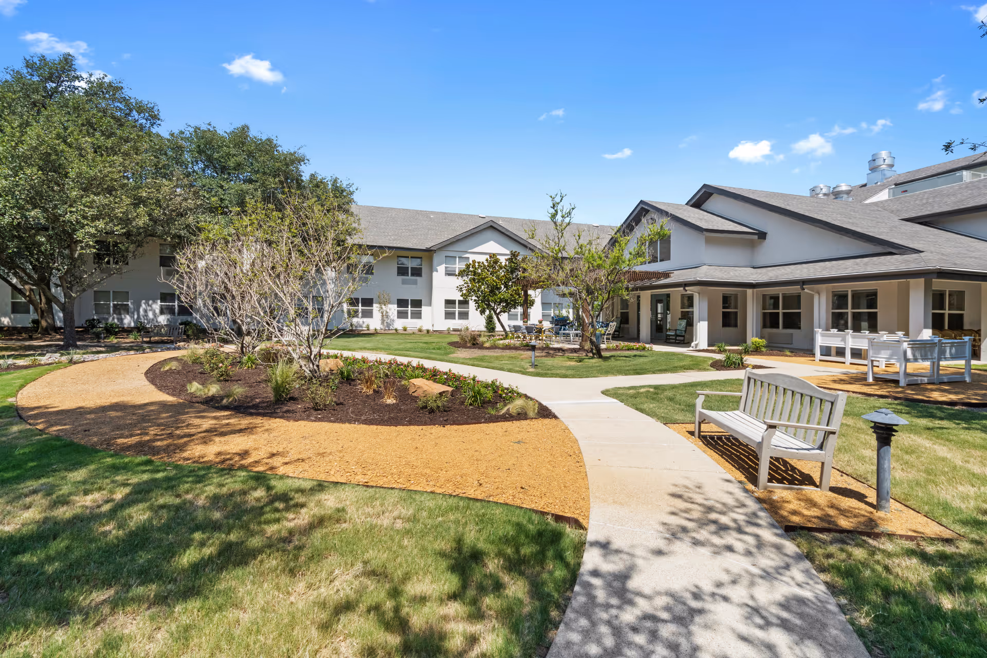 Courtyard and entrance of a senior living facility with pathways, benches, landscaping and a two-story building under a blue sky.