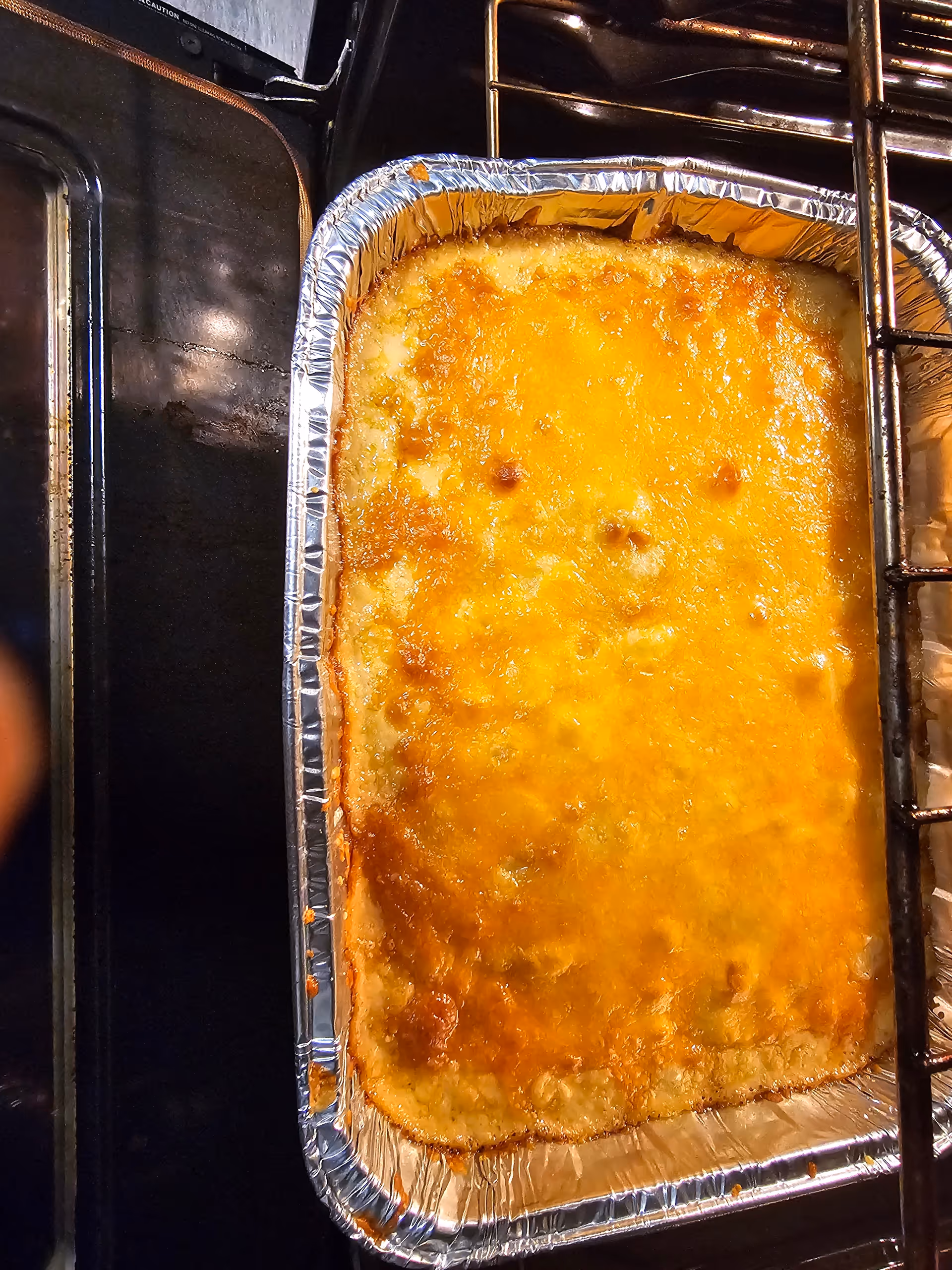 A close-up view of a baked dish with a golden-brown melted cheese topping in a rectangular aluminum foil tray inside an oven.