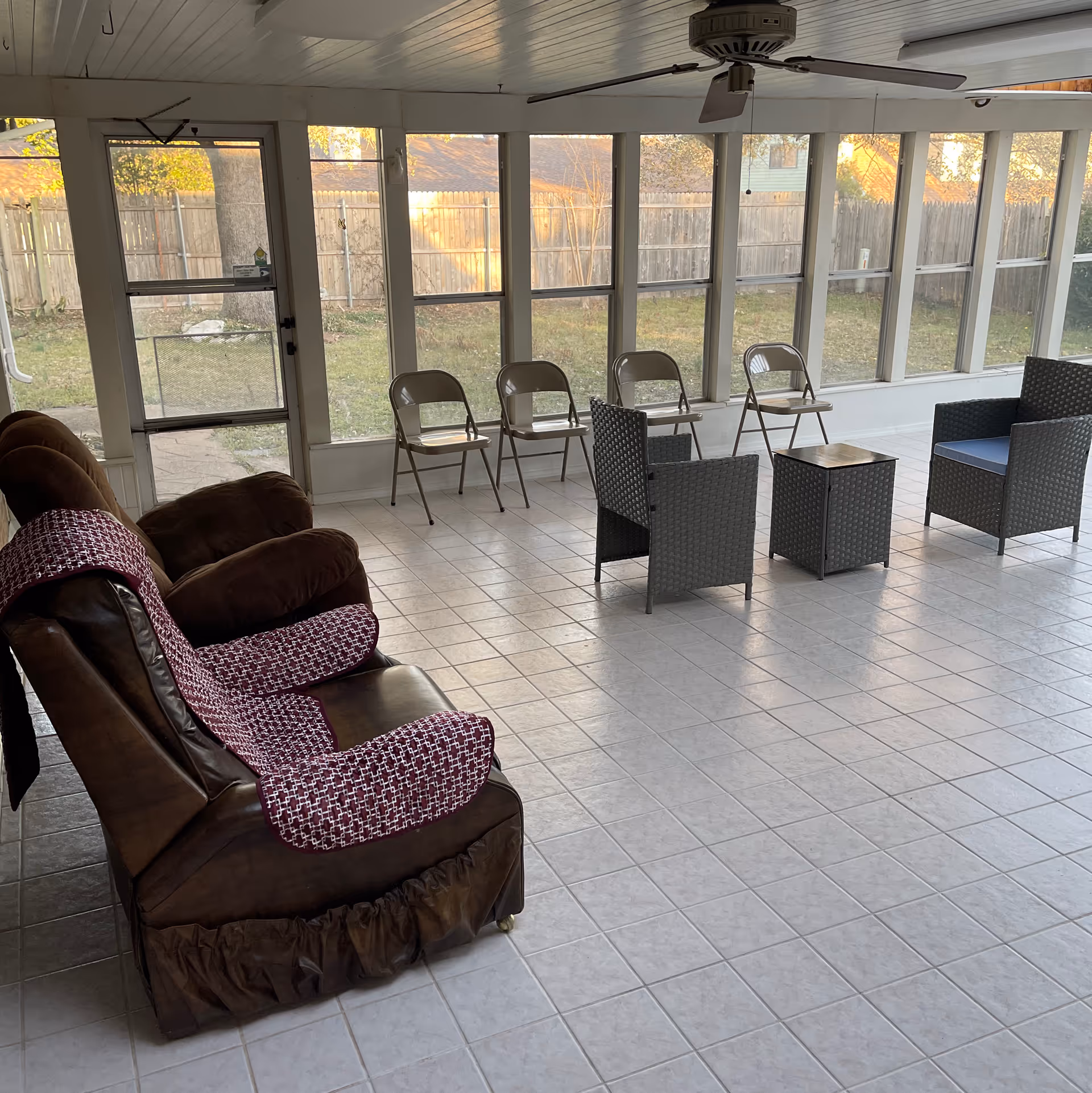 A sunroom with large windows overlooking a fenced backyard. The room has tiled flooring and contains two brown recliner chairs with patterned covers, four metal folding chairs lined up against the windows, two wicker armchairs with blue cushions, and a small wicker table between the armchairs. A ceiling fan is mounted on the white paneled ceiling.