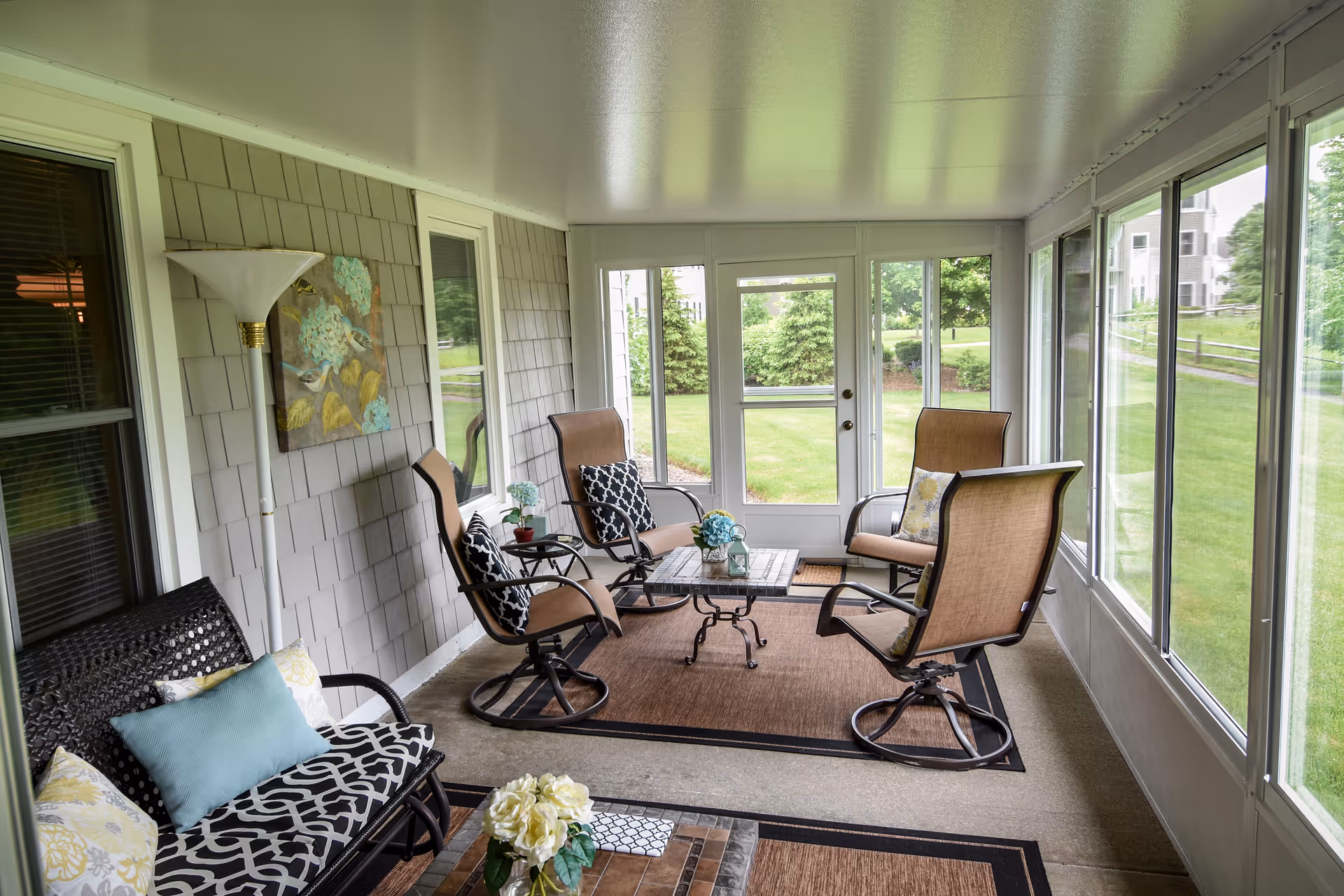A bright enclosed porch area with large windows overlooking a green lawn. The space is furnished with four cushioned swivel chairs around a small square table on a brown rug, and a black wicker bench with patterned cushions and pillows. There is a floor lamp and a floral painting on the wall, and decorative flowers on the table and bench.