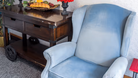 Blue upholstered armchair next to a dark wooden console table topped with a fruit basket and a vase of red flowers.