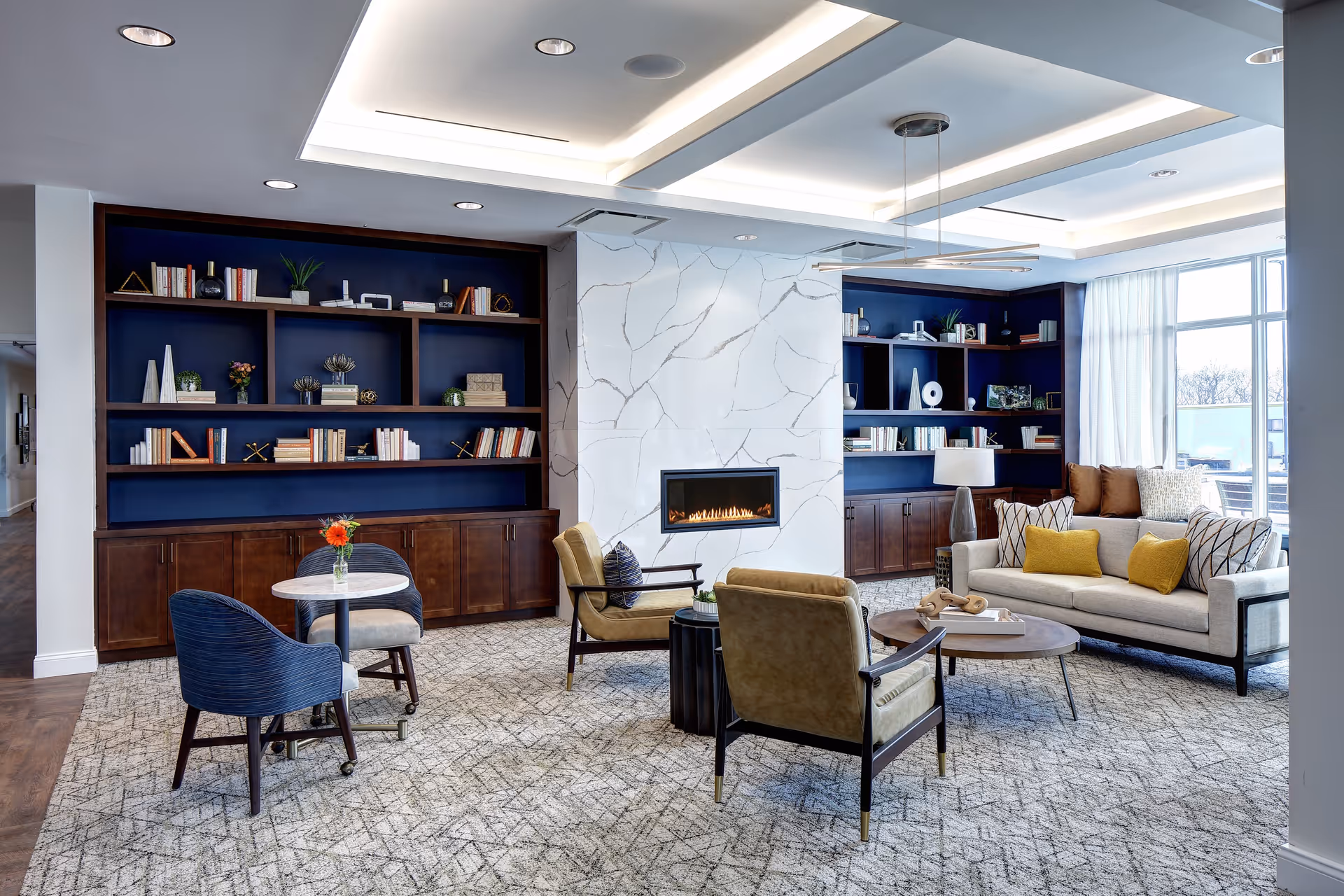 A modern living room area with a light-colored sofa adorned with yellow and patterned pillows, two olive-green armchairs, a round wooden coffee table, and a small round table with two blue chairs. The room features built-in dark wood bookshelves with decorative items and books, a white marble fireplace with a linear gas fire, large windows with sheer curtains, and recessed ceiling lighting.