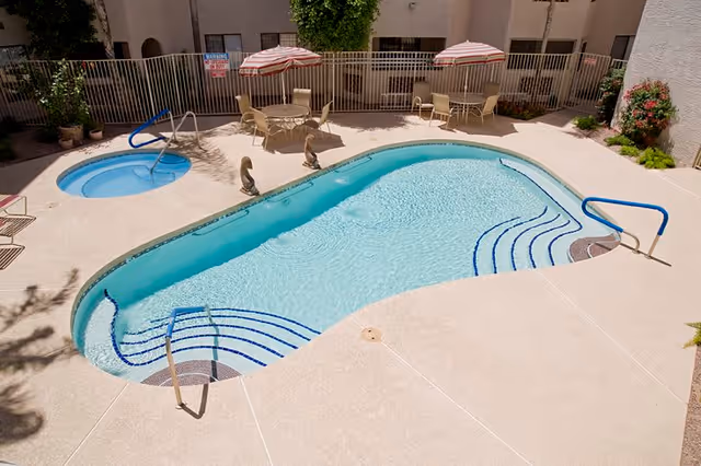 Outdoor swimming pool area with a smaller circular hot tub, surrounded by a beige concrete deck. There are two round tables with umbrellas and chairs near a metal fence enclosing the pool area. Some plants and shrubs are visible along the edges.