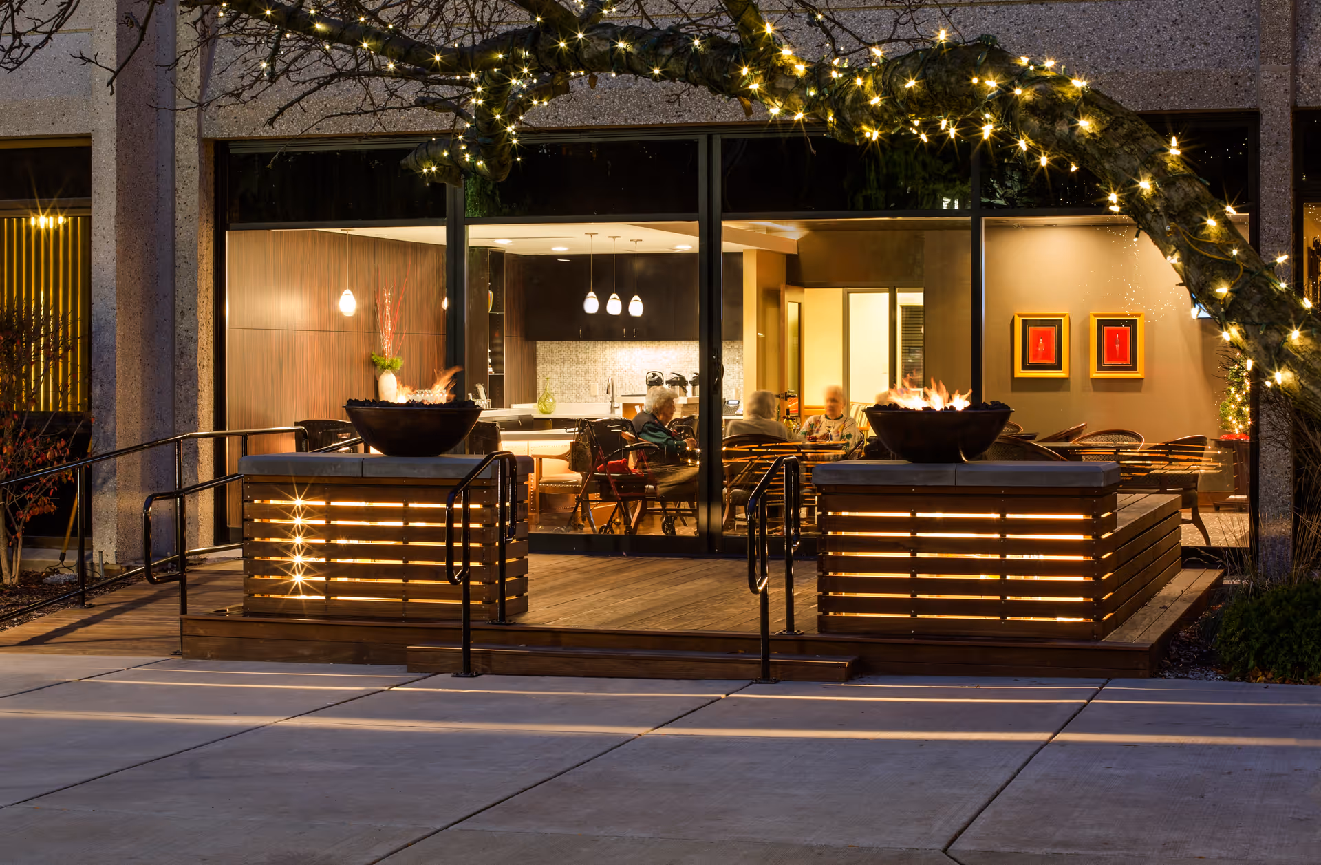 Evening view of a cozy outdoor patio area with two fire pits and string lights wrapped around a tree branch. Through large glass windows, an indoor common area with people sitting and talking around a table is visible, featuring modern lighting and decor.