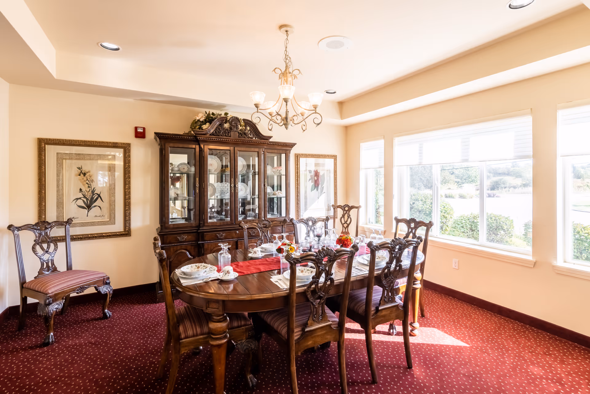 A bright dining room with a large wooden dining table set for a meal, surrounded by eight ornate wooden chairs with striped cushions. Behind the table is a dark wooden china cabinet displaying plates and glassware. The room has large windows letting in natural light, framed artwork on the walls, and a chandelier hanging from the ceiling. The carpet is red with a subtle pattern.