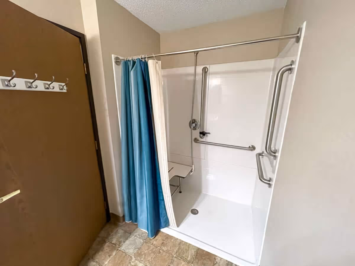 A bathroom shower area with a white walk-in shower equipped with multiple stainless steel grab bars and a foldable shower seat. The shower has a blue and white curtain on a metal rod. To the left, there is a brown door with a white coat rack mounted on it. The floor is tiled with a beige and brown pattern.