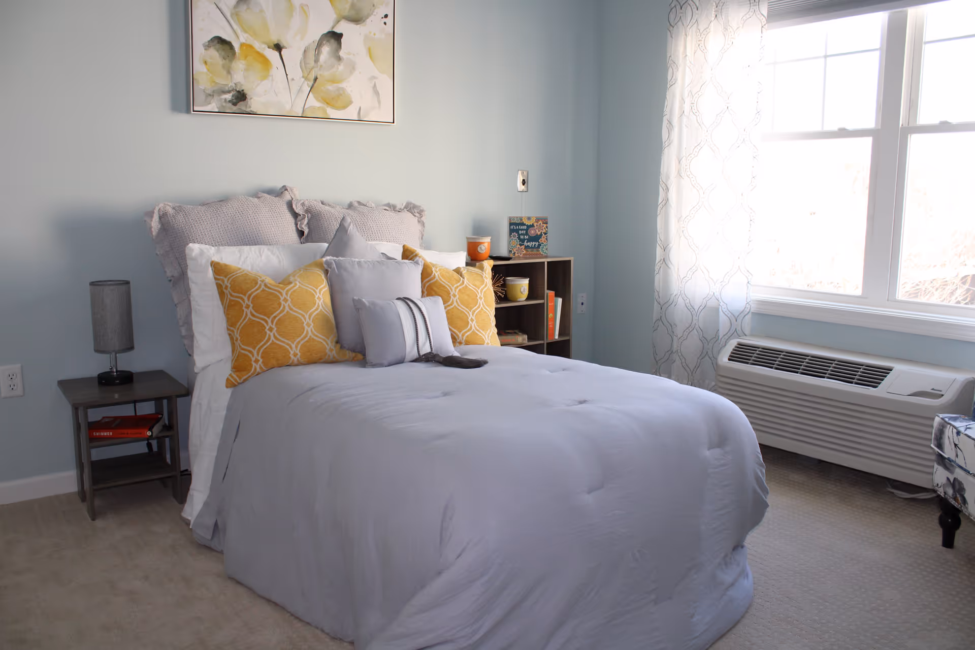 A cozy bedroom with a neatly made bed featuring light gray bedding and multiple pillows in gray and yellow patterns. There is a small nightstand with a lamp and books on the left side of the bed, and a small shelving unit with decorative items and books on the right side. A large window with patterned curtains allows natural light to fill the room. A floral painting hangs on the light blue wall above the bed.