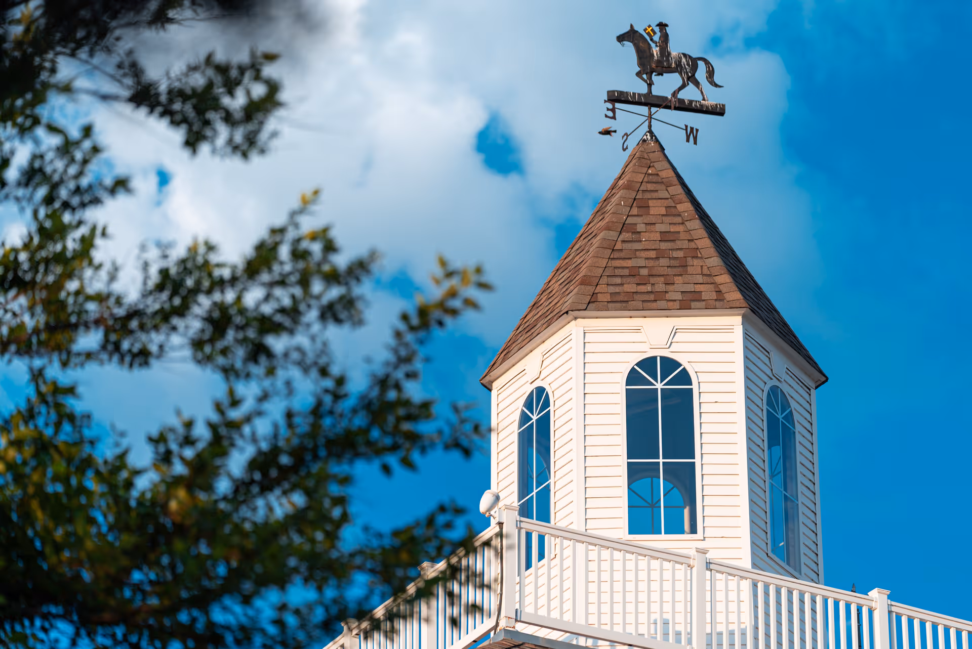 Close-up view of a white tower with arched windows and a brown shingled roof topped with a weather vane featuring a horse and rider, set against a blue sky with some clouds and partially framed by tree branches.