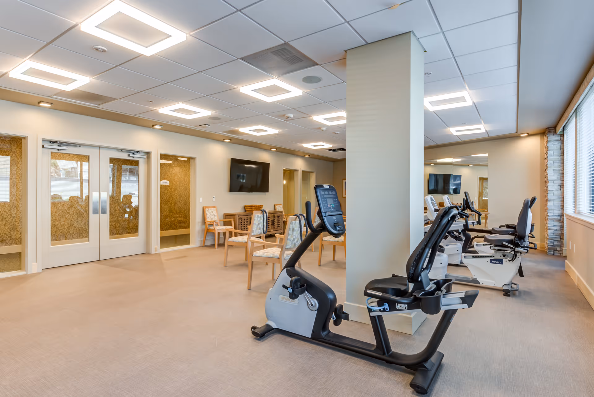 A well-lit exercise room with stationary recumbent bikes lined up facing a wall-mounted TV. The room has beige walls, a large mirror, several chairs, and double doors with glass panels. The ceiling features modern square light fixtures.
