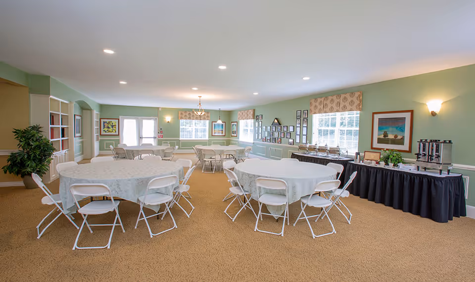 A spacious dining room with round tables covered in white tablecloths and surrounded by white folding chairs. The room has light green walls, beige carpet, and several windows letting in natural light. There is a buffet table along one wall with coffee urns and chafing dishes, and framed artwork and photos decorate the walls.