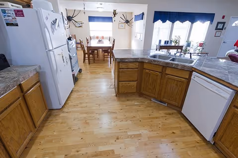 A bright kitchen with wooden cabinets and a light wood floor. The kitchen features a white refrigerator, a dishwasher, a double sink, and a countertop with a marble-like surface. In the background, there is a dining area with a wooden table and chairs, two large windows with blue valances, and wall decorations including two large black spider ornaments.